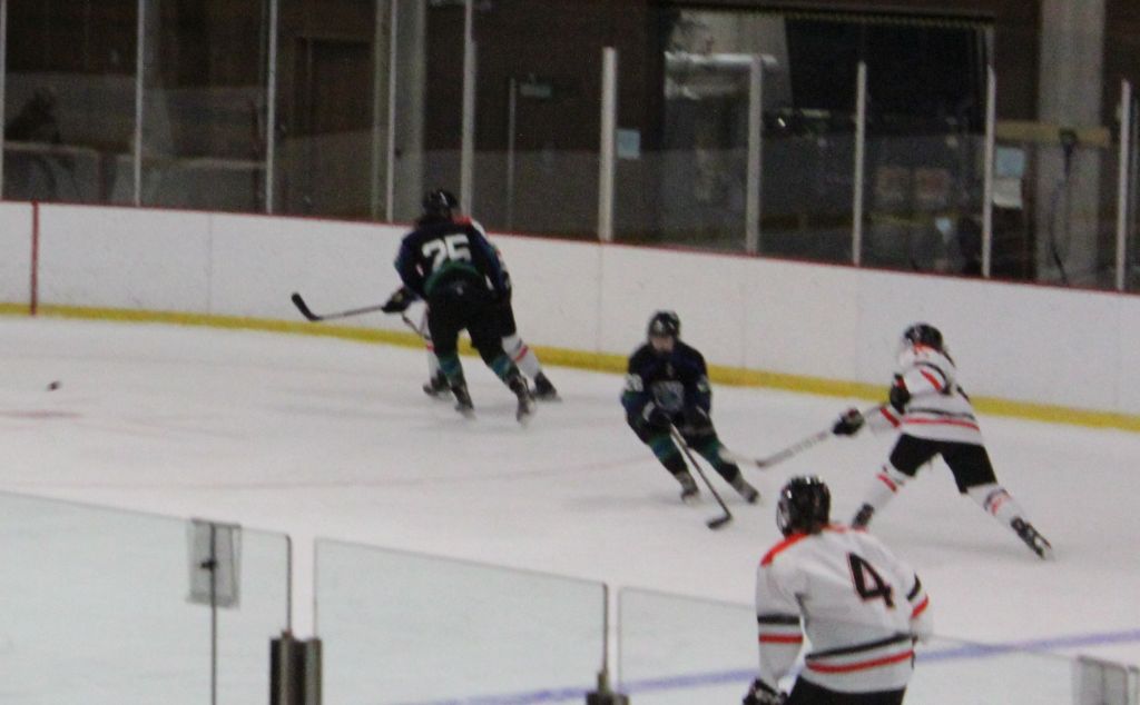 A Wesleyan player in White, red and black shoots the puck as a Salve Regina player in black or navy blue attempts to stop them. Another Wesleyan player is in the foreground.